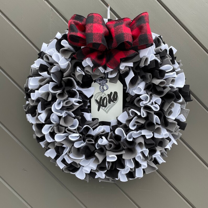May include: A handmade wreath with a mix of black, white, and gray fabric strips. A red and black buffalo plaid bow sits at the top. A tag with "xoxo" and a heart is attached to the wreath.