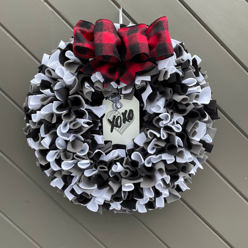 May include: A handmade wreath with a mix of black, white, and gray fabric strips. A red and black buffalo plaid bow sits at the top. A tag with a heart and the word "xoxo" hangs in the center.