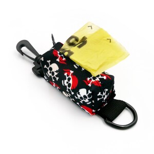 Pode incluir: Dispensador de sacos para dejetos de cães com padrão de caveiras pretas e vermelhas com um saco amarelo parcialmente puxado para fora. O dispensador tem um clipe preto e um anel preto.