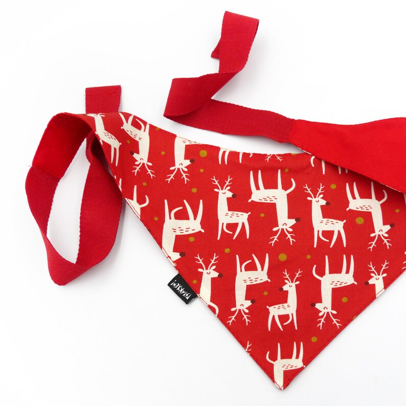 May include: Red and white patterned bandana with a reindeer design. The bandana has a red strap that can be tied around the neck.