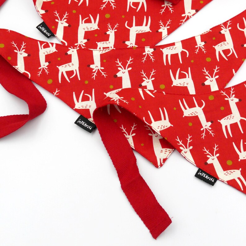May include: Two red fabric dog bandanas with a white reindeer pattern and gold dots. The bandanas have a red ribbon attached and the word "remarkable" printed on the tag.