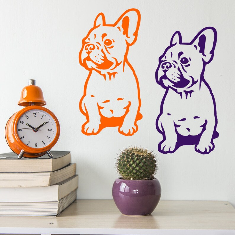 May include: Two silhouette wall decals of French Bulldogs, one in orange and one in purple, on a white wall. The decals are above a stack of books and a small potted cactus.