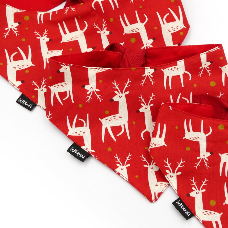 May include: Three red fabric dog bandanas with a white reindeer pattern and gold polka dots. The bandanas have black tags with the text "i-K9-i" printed on them.