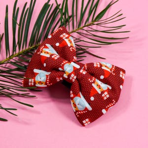 Könnte beinhalten: Eine rote Fliege mit weißem Tupfenmuster und einem sich wiederholenden Muster von Weihnachtsmannfiguren. Die Fliege ist aus Stoff und hat ein festliches Weihnachtsmotiv.
