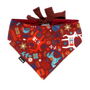 Puede incluir: Un pañuelo rojo con un estampado festivo de inspiración escandinava que presenta renos, pájaros, estrellas y formas geométricas en tonos azules, blancos, naranjas y dorados. El pañuelo tiene una corbata burdeos.