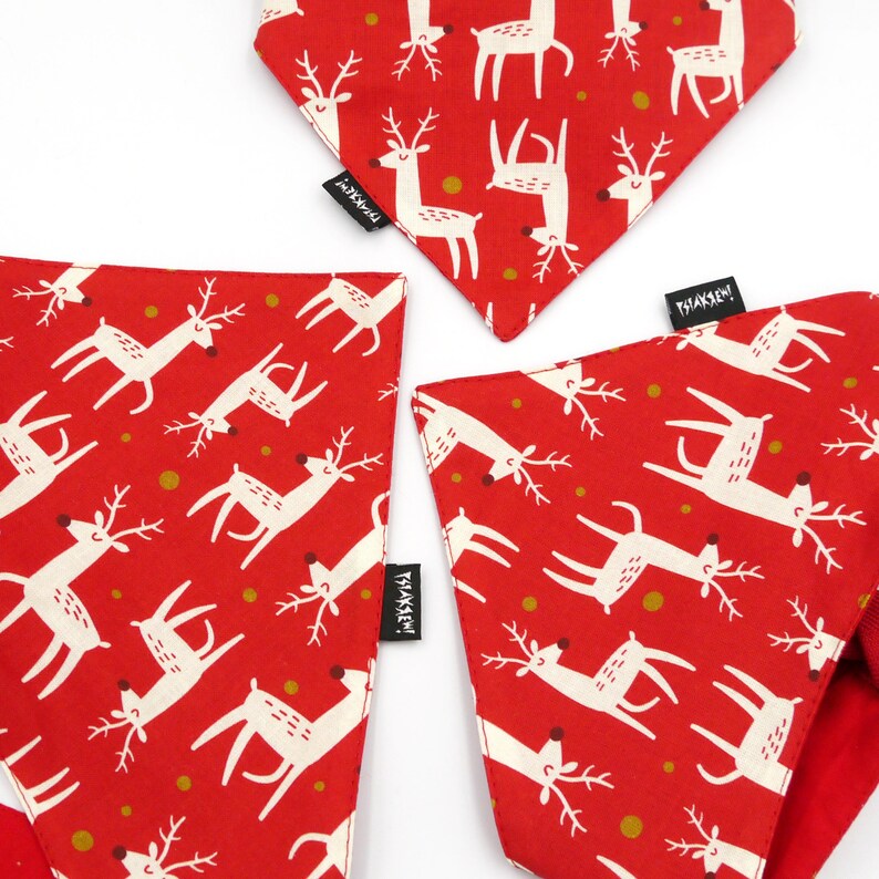 May include: Three red bandana style dog bandanas with a white reindeer pattern and gold dots. The bandanas have a white tag with the text "Tisket" on them.