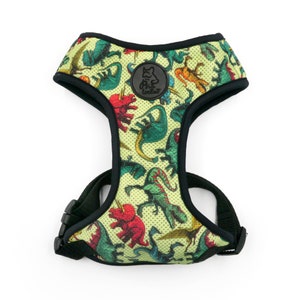 Puede incluir: Un arnés para perro verde con estampado de dinosaurios. El arnés tiene correas negras y una hebilla negra. El arnés tiene un logotipo que dice "Pup Luxe" en él.