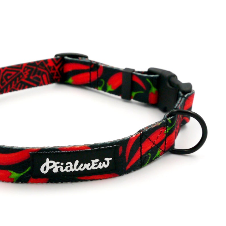 Puede incluir: Un collar de perro negro con estampado de chiles rojos y una hebilla negra. El collar tiene una etiqueta negra con el texto "Primal Pet" en blanco.