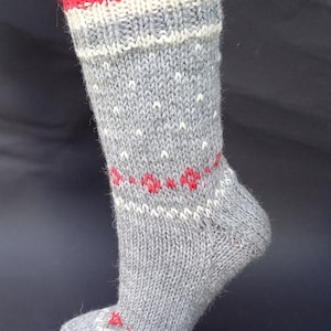 Peut inclure: Une chaussette grise tricotée avec des accents rouges et blancs. Le bord-côte est rayé rouge, blanc et gris. La chaussette présente un motif floral rouge répété et des mouchetures blanches. La chaussette est présentée sur un pied de mannequin.