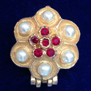 May include: A gold-toned brooch with a flower design. The center of the flower has a cluster of red rhinestones. The flower is surrounded by six white pearls.