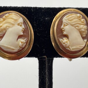 May include: A pair of gold tone cameo earrings featuring a carved woman's profile in white against a brown background.