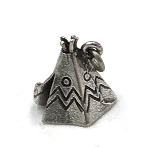 May include: A silver-toned metal charm shaped like a teepee with a conical structure, featuring engraved geometric zigzag patterns and circular motifs on its exterior surface, topped with small poles and including a circular jump ring attachment.