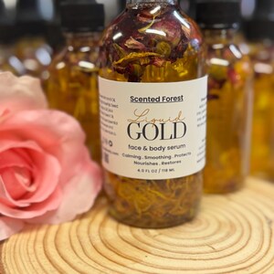 May include: A clear glass bottle of face and body serum, labeled "Liquid Gold" by Scented Forest. The bottle contains visible botanicals and is topped with a black dropper. The label indicates 4.0 fl oz / 118 ml. A pink rose and wooden surface are in the foreground.