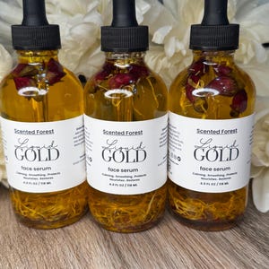 May include: Three glass bottles of face serum with black droppers. The bottles are filled with a golden liquid and contain dried rose petals. Each bottle has a white label with the text "Liquid Gold face serum" and "Scented Forest". Each bottle contains 4.0 fl oz / 118 ml.