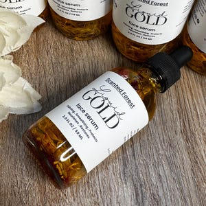 May include: Several bottles of Scented Forest Liquid Gold face serum are arranged on a wooden surface. The bottles are clear, filled with a golden liquid, and have white labels with black text. The labels read "Liquid Gold face serum" and include the volume: 2.0 FL OZ / 59 ML.