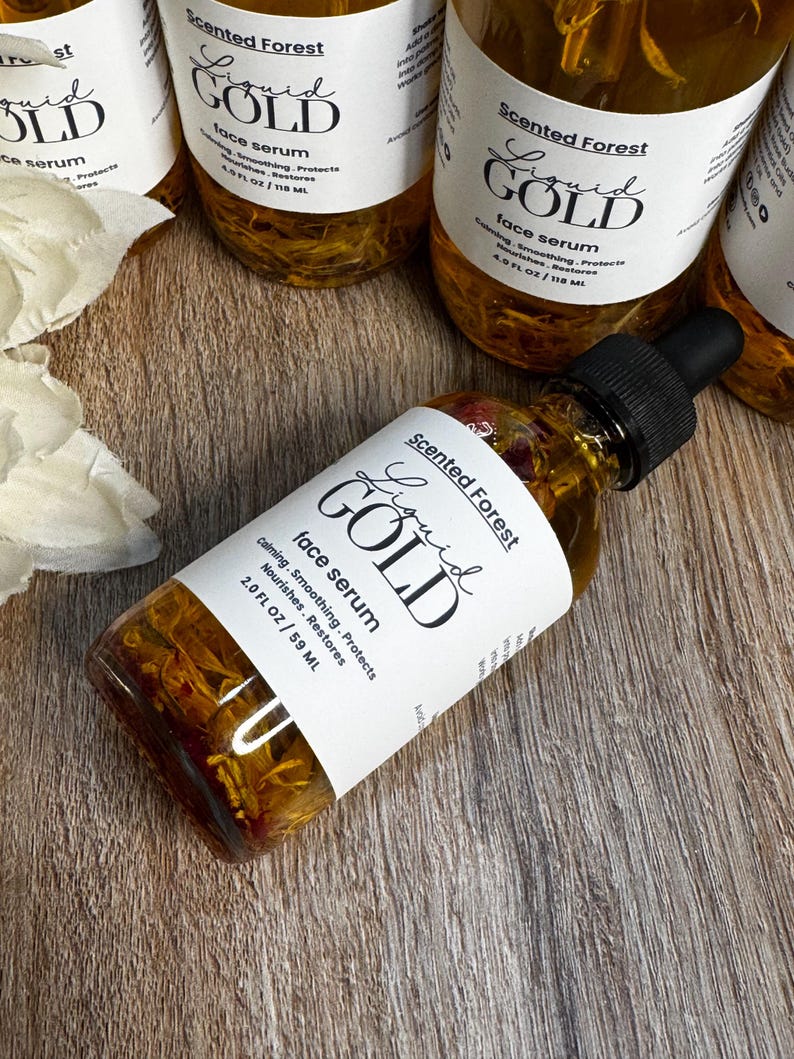 May include: Several bottles of Scented Forest Liquid Gold face serum are arranged on a wooden surface. The bottles are clear, filled with a golden liquid, and have white labels with black text. The labels read "Liquid Gold face serum" and include the volume: 2.0 FL OZ / 59 ML.