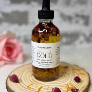 May include: A clear glass bottle of "Liquid Gold" face and body serum, with a black dropper top. The bottle contains a yellow liquid with dried rose petals and other botanicals. The bottle sits on a wooden slice with dried rose buds and petals.