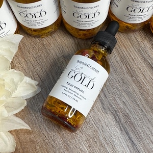 May include: Several bottles of Scented Forest Liquid Gold face serum are displayed. The bottles are filled with a golden liquid and contain floral elements. Each bottle has a white label with black text. The label reads "Liquid Gold face serum".