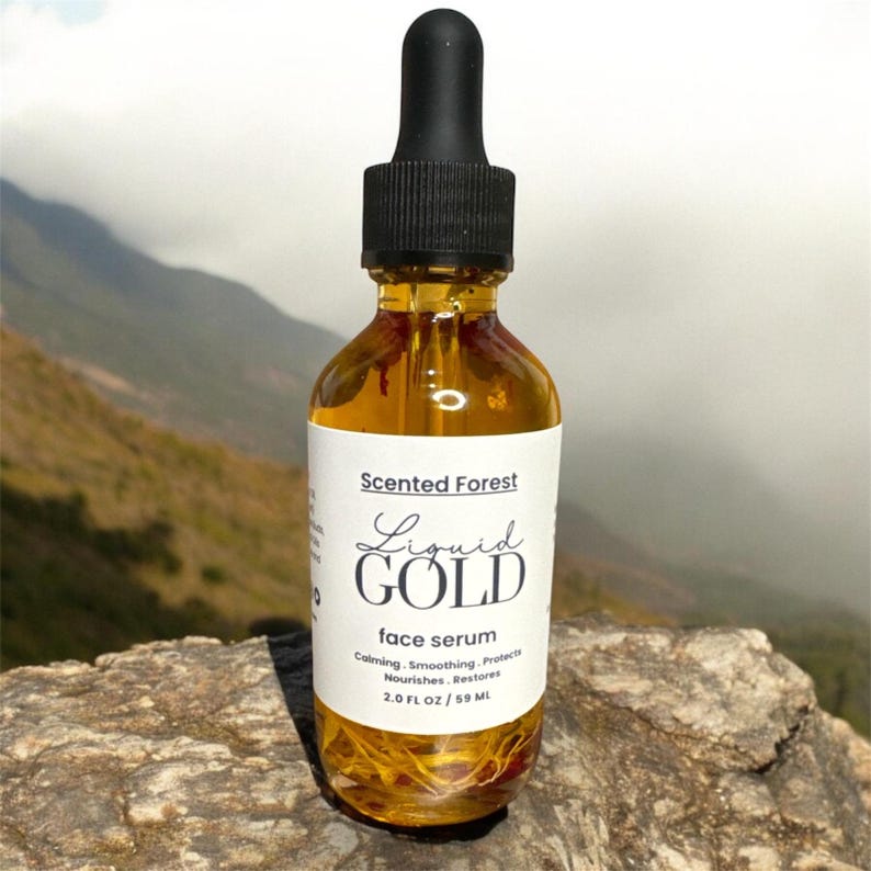 May include: A clear glass bottle of face serum with a black dropper. The label reads "Scented Forest Liquid GOLD face serum". The bottle contains a golden liquid and is 2.0 fl oz / 59 ml. The background shows a mountain range.
