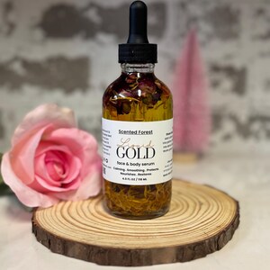 May include: A clear glass bottle of face and body serum with a black dropper, labeled "Liquid Gold" by Scented Forest. The bottle contains visible botanicals and sits on a wooden slice with a pink rose in the foreground. The serum is 4.0 fl oz / 118 ml.