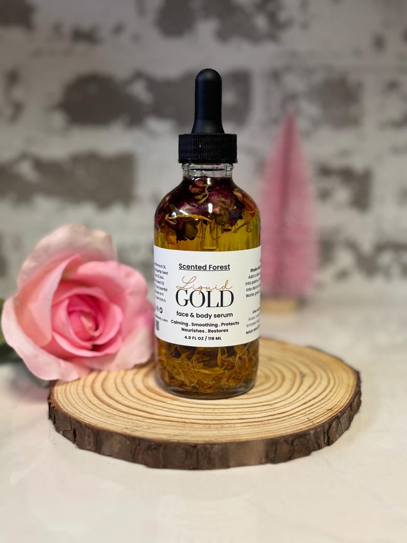 May include: A clear glass bottle of face and body serum with a black dropper, labeled "Liquid Gold" by Scented Forest. The bottle contains visible botanicals and sits on a wooden slice with a pink rose in the foreground. The serum is 4.0 fl oz / 118 ml.
