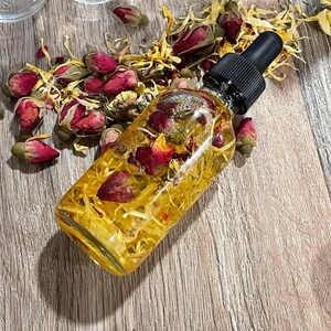 May include: A clear glass bottle filled with a yellow liquid and dried rose petals and buds. The bottle has a black dropper top. Dried yellow flowers and pink rose buds are scattered around the bottle on a wooden surface.