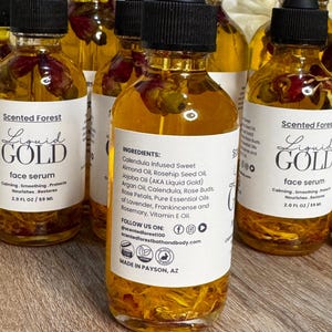 May include: A collection of glass bottles filled with a golden liquid, each topped with a black dropper. The bottles have white labels with the text "Liquid Gold face serum" and ingredient information. The liquid contains rose petals.