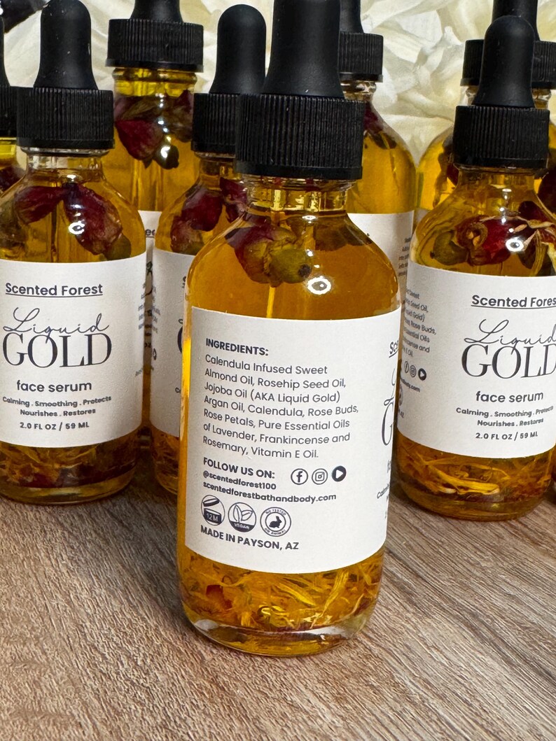 May include: A collection of glass bottles filled with a golden liquid, each topped with a black dropper. The bottles have white labels with the text "Liquid Gold face serum" and ingredient information. The liquid contains rose petals.