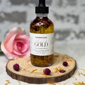May include: A clear glass bottle of "Liquid Gold" face and body serum with a black dropper. The bottle contains dried flowers and is set on a wooden slice with dried flower buds and petals. The label reads "Scented Forest".