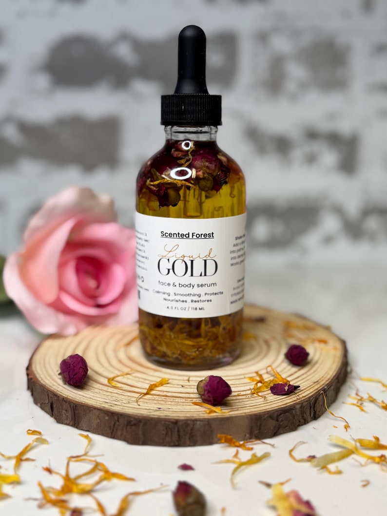 May include: A clear glass bottle of "Liquid Gold" face and body serum with a black dropper. The bottle contains dried flowers and is set on a wooden slice with dried flower buds and petals. The label reads "Scented Forest".