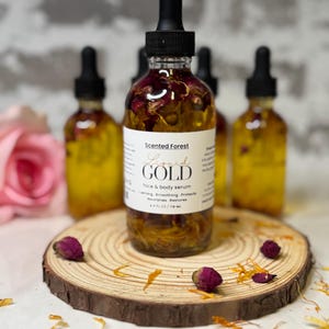 May include: A collection of amber-colored glass bottles filled with face and body serum, each topped with a black dropper. The bottles are adorned with dried flower petals and sit on a wooden slice. The label reads "Liquid Gold".