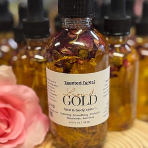 May include: A clear glass bottle of "Liquid Gold" face and body serum, filled with botanicals. The label reads "Scented Forest" and lists benefits. The bottle has a black dropper top and contains 4.0 fl oz / 118 ml of product.