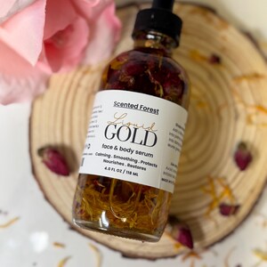 May include: A clear glass bottle of "Liquid Gold" face and body serum with a black dropper. The bottle contains dried flowers and petals. The label reads "Scented Forest" and lists the serum's benefits. The bottle is 4.0 fl oz / 118 ml.