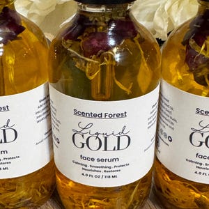 May include: Three clear glass bottles of face serum with black caps. The bottles contain a golden liquid with dried flowers. Each bottle has a white label with the text "Liquid Gold face serum" and "Scented Forest". Each bottle contains 4.0 FL OZ / 118 ML.