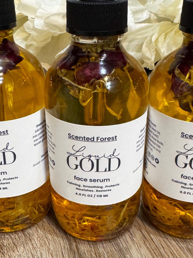 May include: Three clear glass bottles of face serum with black caps. The bottles contain a golden liquid with dried flowers. Each bottle has a white label with the text "Liquid Gold face serum" and "Scented Forest". Each bottle contains 4.0 FL OZ / 118 ML.