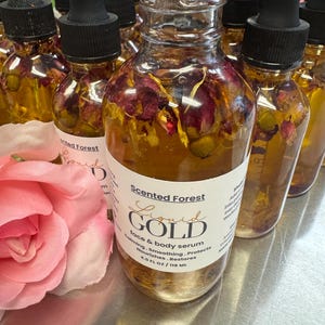 May include: Clear glass bottles filled with golden liquid and dried flowers. Each bottle has a black dropper and a label that reads "Scented Forest Liquid Gold face & body serum 4.0 FL OZ / 118 ML". A pink rose is in the foreground.