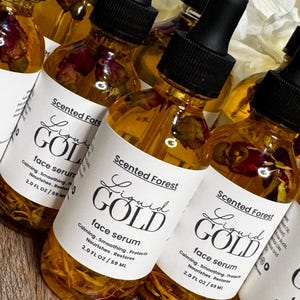 May include: Several amber-colored glass bottles of face serum with black droppers are displayed. Each bottle has a white label with the text "Liquid Gold" and "face serum". The bottles contain dried flower petals.