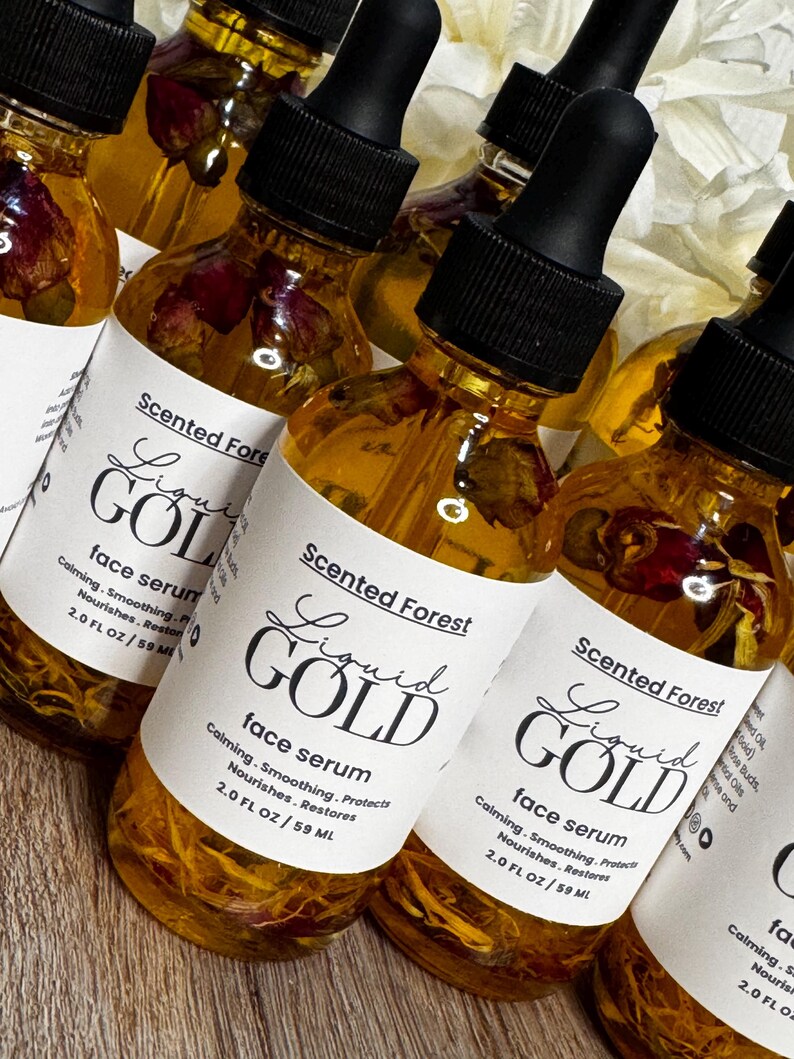 May include: Several amber-colored glass bottles of face serum with black droppers are displayed. Each bottle has a white label with the text "Liquid Gold" and "face serum". The bottles contain dried flower petals.