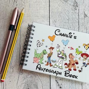 Op de afbeelding: Een wit spiraalgebonden autographboek met een kleurrijke afbeelding van Toy Story-personages zoals Woody, Buzz Lightyear, Jessie, Rex, Bullseye en Hamm, met de tekst Charlie's Autograph Book, vergezeld door een metallic roze pen en een geel potlood.