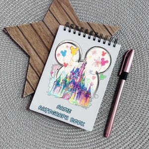 Op de afbeelding: Een spiraalgebonden handtekeningenboek met een kleurrijk kasteelontwerp en de woorden "NAME AUTOGRAPH BOOK". Een roségouden pen ligt naast het boek. Het boek ligt op een grijze placemat met een houten ster.