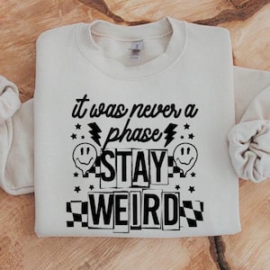 May include: A cream-coloured sweatshirt featuring the black text "it was never a phase", lightning bolts, stars, and smiley faces. The word "STAY" is in bold, and "WEIRD" is in a chequered pattern.