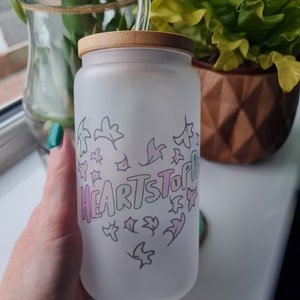Op de afbeelding: Een witte, matte glazen beker met een bamboe deksel en een heldere glazen rietje. De beker heeft een roze en grijze afbeelding van een hart met de woorden "HEARTSTOPPER" erin.