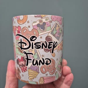 May include: A light blue ceramic mug with a Disney-themed pattern. The mug features a repeating design of Mickey Mouse-themed images, including a camera, pretzels, and cupcakes. The words "Disney Fund" are written in black script across the front.