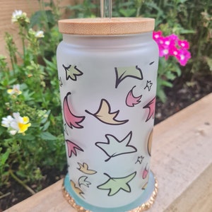 Op de afbeelding: Een frosted glazen beker met een bamboe deksel en een helder glazen rietje. De beker is versierd met kleurrijke bladeren. De basis heeft een gouden glitterrand. De beker is ongeveer 15 cm hoog.