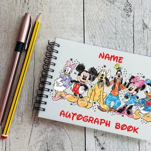 May include: A white spiral-bound autograph book with a vibrant illustration of Disney characters, including Mickey Mouse and Donald Duck. The cover reads "NAME" and "AUTOGRAPH BOOK" in red. A pink pen and a yellow pencil are placed beside the book.