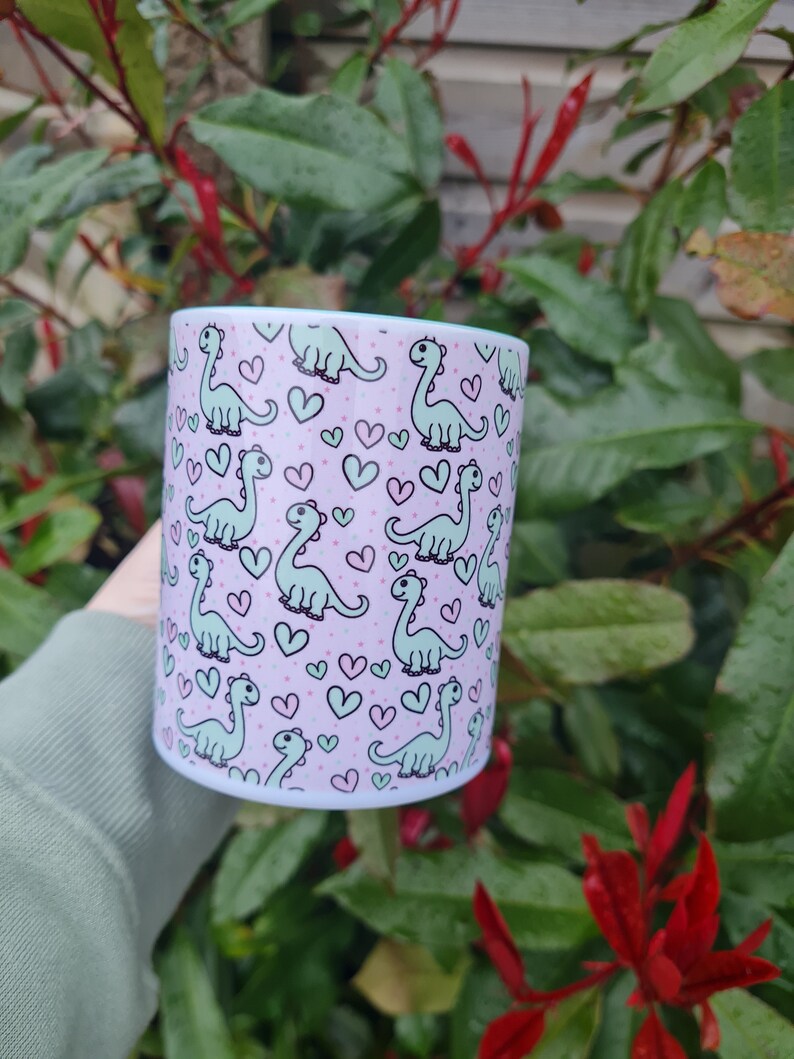 Op de afbeelding: Een witte keramische mok met een roze achtergrond en een herhalend patroon van groene dinosaurussen en roze harten.