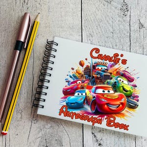 Op de afbeelding: Een wit spiraalgebonden autografenboek met een kleurrijke illustratie van tekenfilmauto's en de tekst "Charlie's Autograph Book". Een roségouden pen en een gele potlood liggen naast het boek.