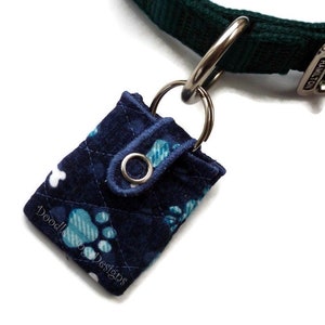 May include: A blue quilted dog poop bag holder with a paw print design. The holder is attached to a green dog collar with a silver buckle. The text "Doodle Dog Designs" is embroidered on the holder.