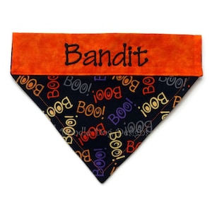 Puede incluir: Un pa&ntilde;uelo para perro con tem&aacute;tica de Halloween con el nombre "Bandit" bordado en negro sobre un panel naranja. La secci&oacute;n principal es negra con "BOO!" en varios colores.