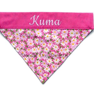 May include: Pink dog bandana with a white daisy print and a pink polka dot border. The bandana is personalized with the name "Kuma" in white embroidery.